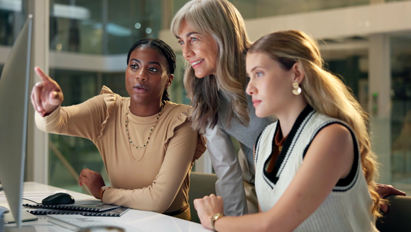 Three women engaged in a discussion while looking at a computer screen in a modern office environment showcasing teamwork and collaboration in a professional setting.